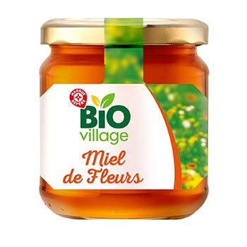A glass jar of BIO village Miel de Fleur (blossom honey) with a gold metal lid and a white label featuring green and orange accents, isolated on a white background.